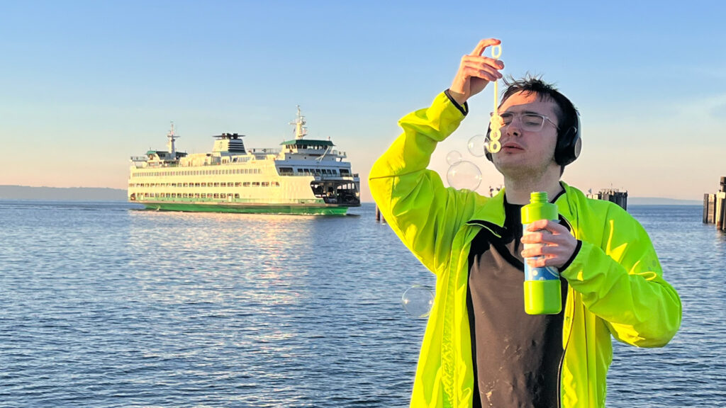 Autistic teen with apraxia blows bubbles at the waterfront while wearing a neon yellow jacket and headphones; a Washington State ferry passes in the background during golden hour, capturing a peaceful sensory moment.
