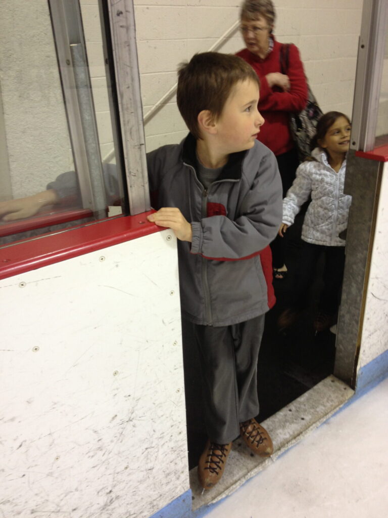 Stone, age 7, standing at the threshold of an ice rink in rental skates, looking out with curiosity and hesitation.