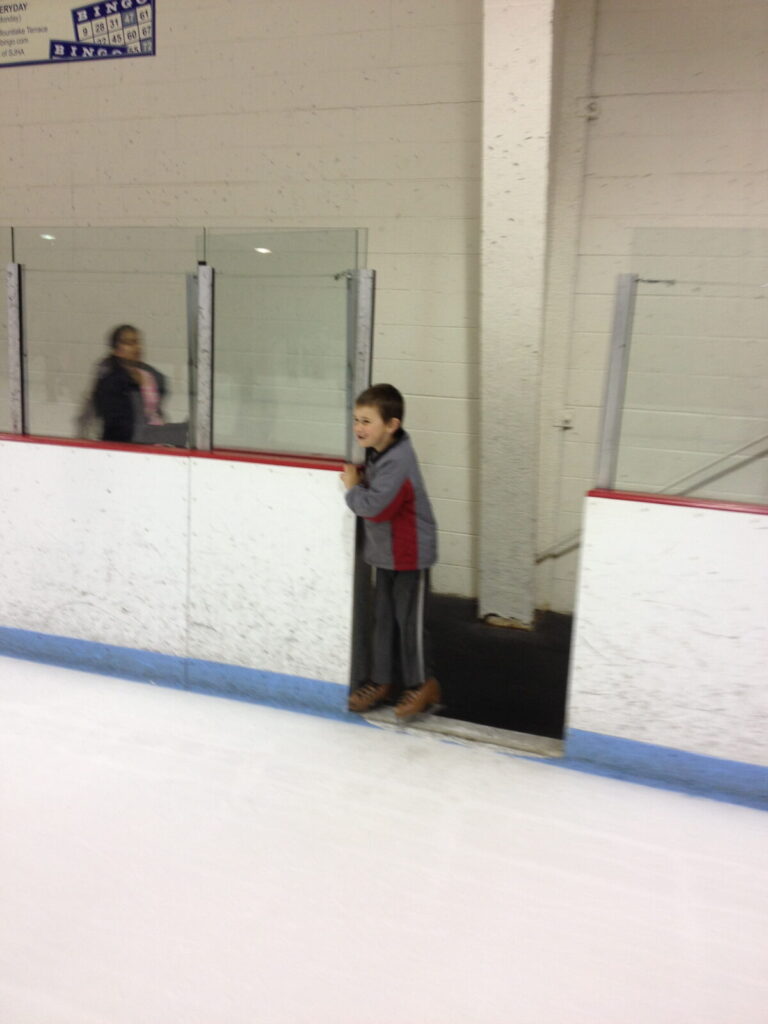 Stone smiling while standing at the rink entrance, in the same skates, beaming with joy during his first successful skating experience.