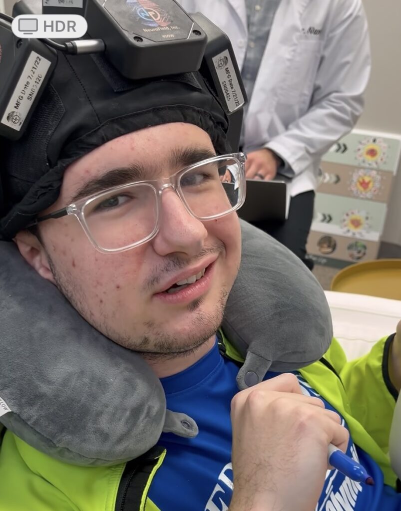 Stone smiles while wearing a neurotherapy cap during a Brain Enhancement Therapy session. He’s holding a pen and wearing a travel pillow for support, with a technician visible in the background. His expression is relaxed, confident, and full of quiet pride.