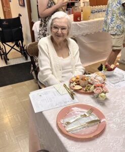 Diane Kaufer, a smiling 90-year-old woman with silver hair and glasses, seated at a decorated table during her birthday celebration. She wears a patterned blouse and holds a bouquet of pink and white flowers, with warm lighting and joyful energy in the background.